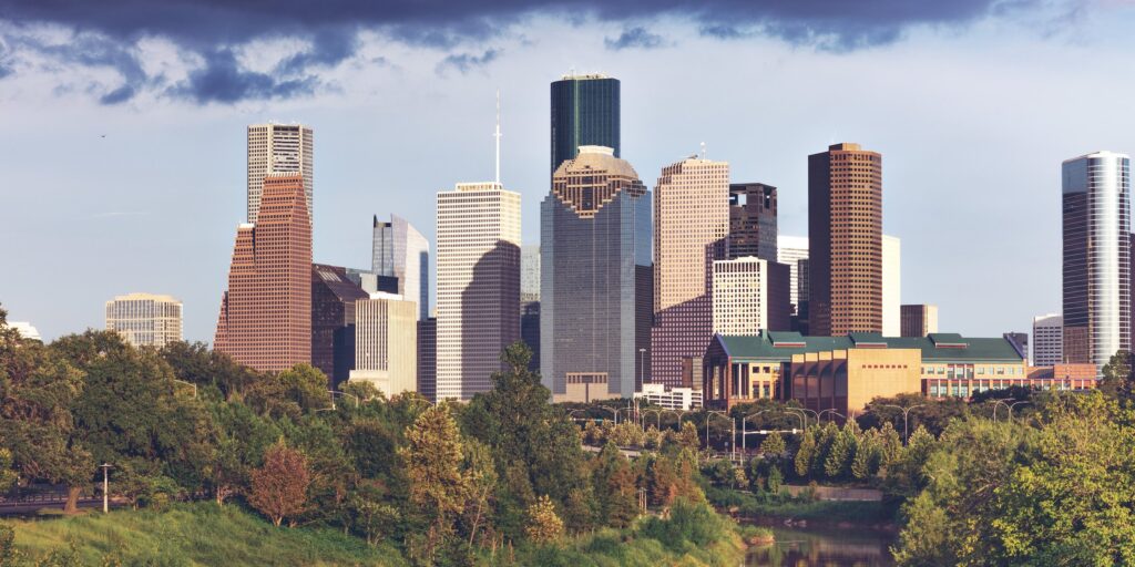 The downtown Houston skyline seen from the west over Buffalo Bayou, with trees in the foreground under a cloudy sky