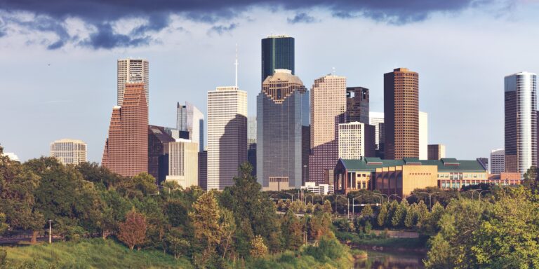 The downtown Houston skyline seen from the west over Buffalo Bayou, with trees in the foreground under a cloudy sky